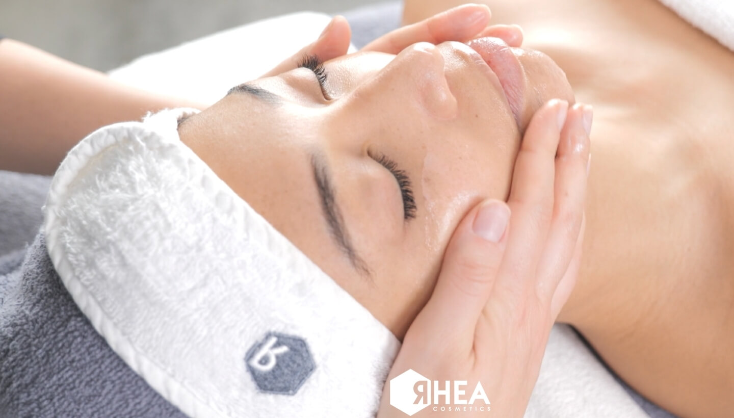 Massaggio del viso con mani, persona con asciugamano e logo Rhea Cosmetics.
