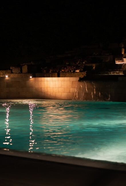 Piscina illuminata di notte con acqua che scorre su una parete rocciosa.