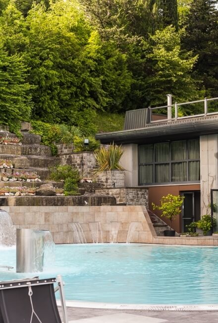 Piscina con cascate, edificio moderno e vegetazione lussureggiante circostante.
