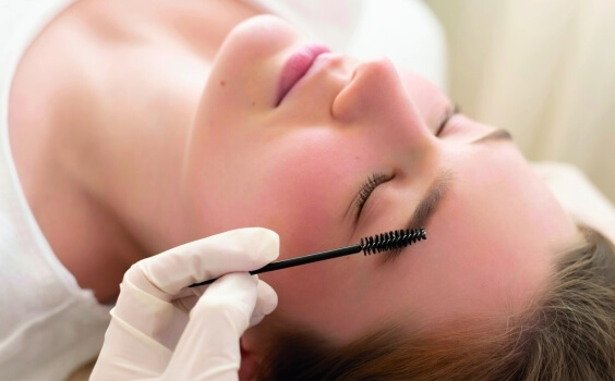 Person with closed eyes, gloved hand applying mascara to eyebrow. Person mit geschlossenen Augen, Handschuh hält Mascara für Augenbraue.