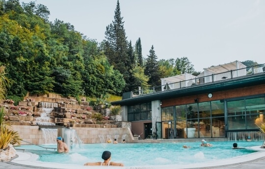 Outdoor pool with waterfall, people swimming, modern building, and greenery.