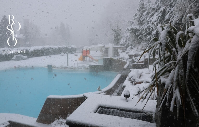 Piscina all'aperto con acqua blu sotto la neve, scivoli visibili.