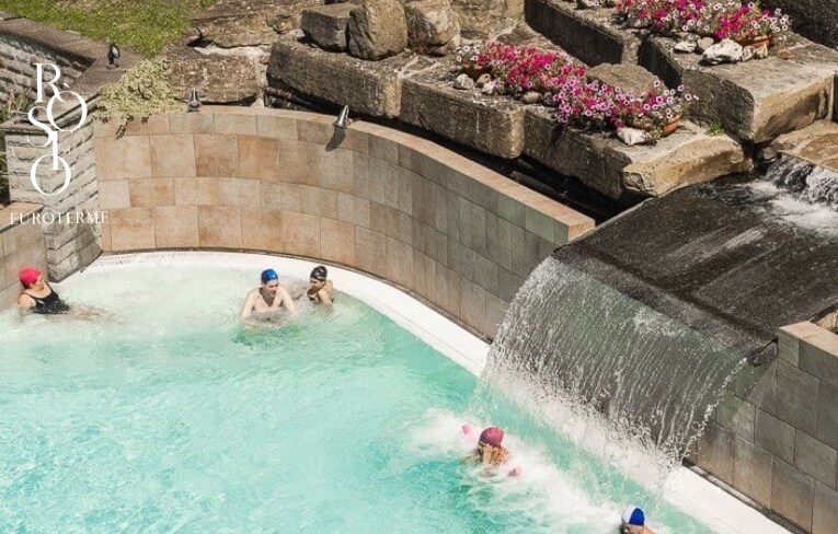 Piscina con cascata, persone, fiori rosa e testo "ROSO EUROTERME".