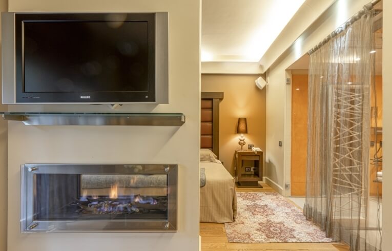 Camera d'hotel con TV, camino, letto e doccia in vetro.