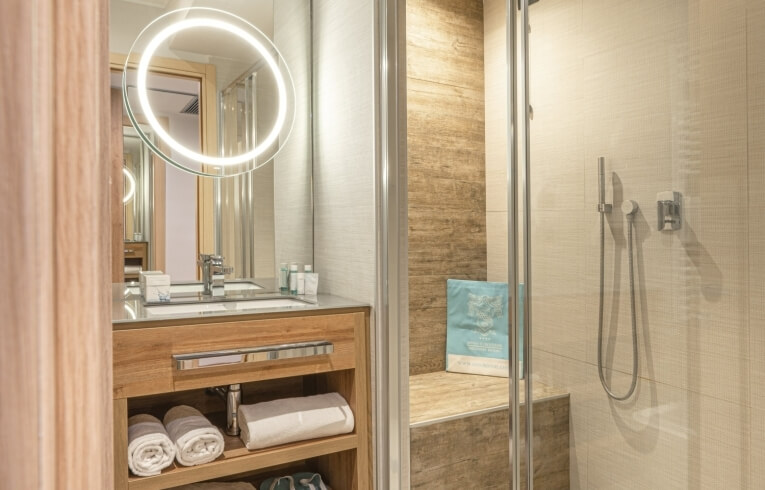 Hotel bathroom with illuminated mirror, shower enclosure, and towel shelves.