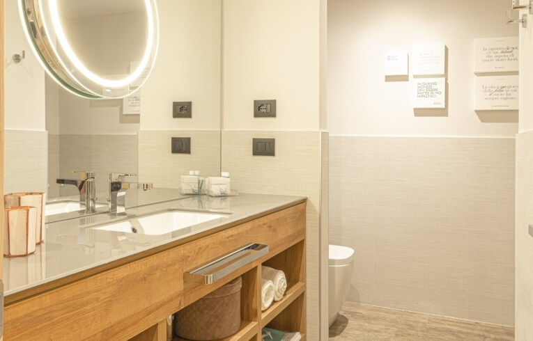 Hotelbadezimmer mit Doppelwaschtisch, ovalem Spiegel und Holzregal.