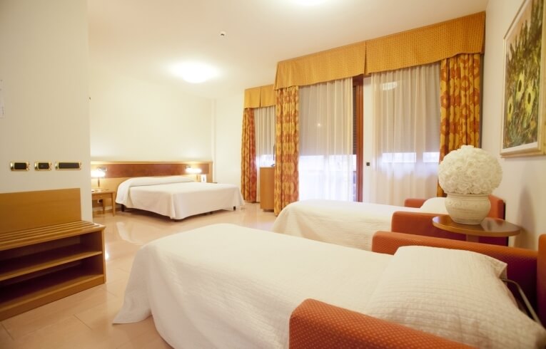 Hotel room with three beds, orange chairs, and patterned curtains.
