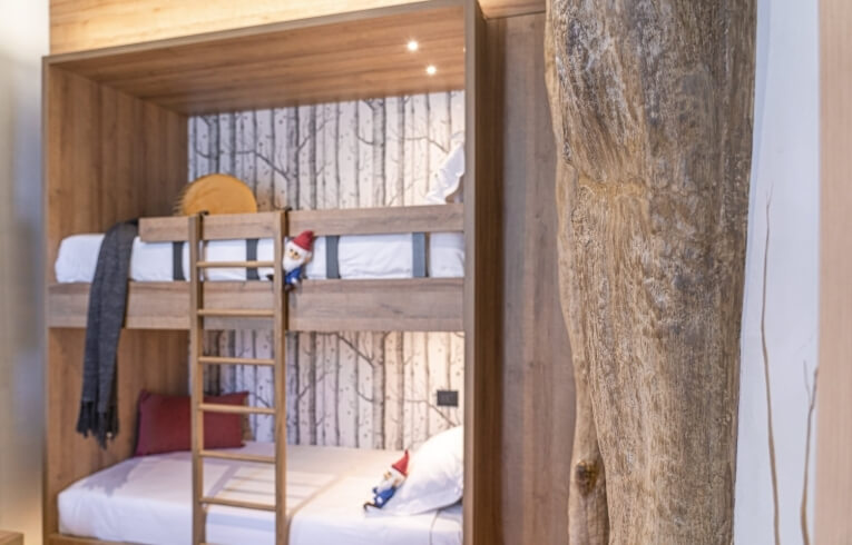 Wooden bunk bed with grey blanket, gnome pillow, and tree wallpaper.