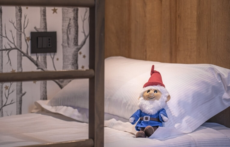 Stuffed gnome on a bed with striped bedding and tree-patterned wallpaper.