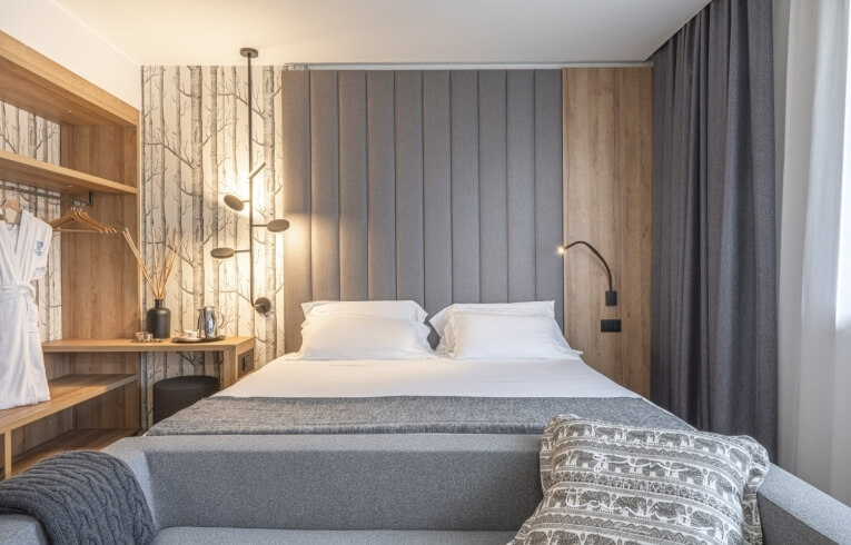 Hotelzimmer mit Bett, grauem Sofa, Teekanne und hängendem Bademantel.