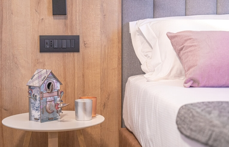 Bedside table with birdhouse, containers, and light switch on wooden wall.