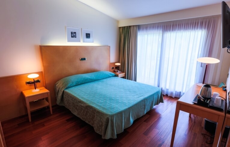 Camera d'hotel con letto teal, scrivania e finestre con tende.