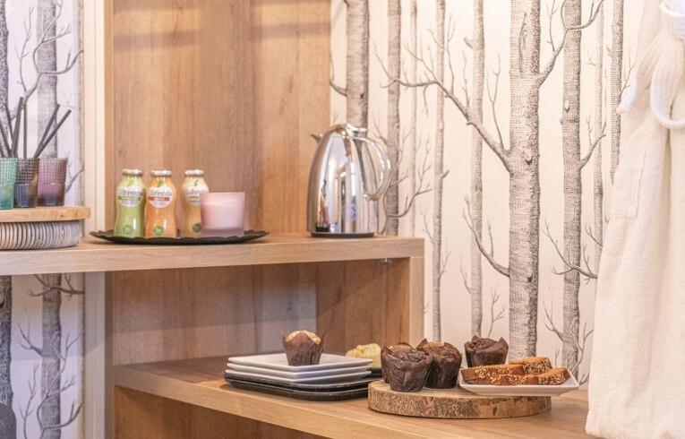 Wooden shelf with juice bottles, candle, thermos, plates, muffins, and bread.