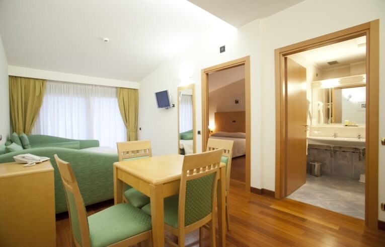 Camera d'hotel con divano verde, tavolo, TV e porte per bagno e camera da letto.