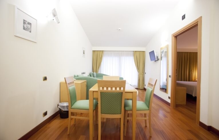 Camera d'hotel con tavolo, divano, TV e porta verso la camera da letto.