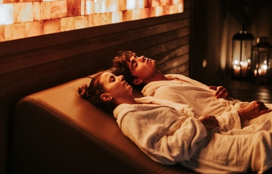 Due persone in accappatoi su lettini, con parete di mattoni di sale rosa illuminati.