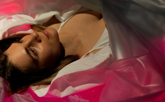 Donna sdraiata in una maglietta bianca, circondata da materiali traslucidi illuminati di rosa e rosso.