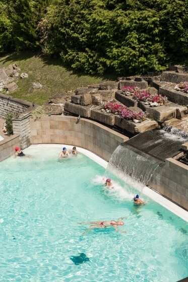 Piscina con persone, cascata, fiori rosa e alberi sullo sfondo.