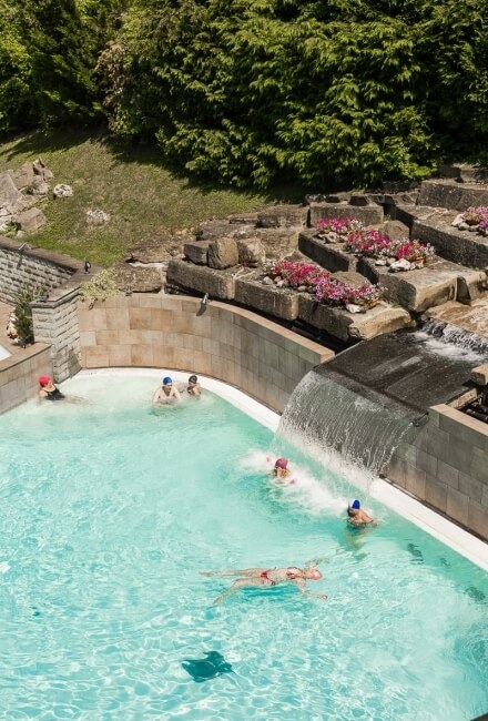 Piscina all'aperto con cascata, persone che nuotano e vegetazione.