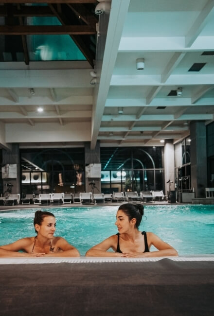 Due donne in una piscina interna con acqua turchese e finestre grandi.