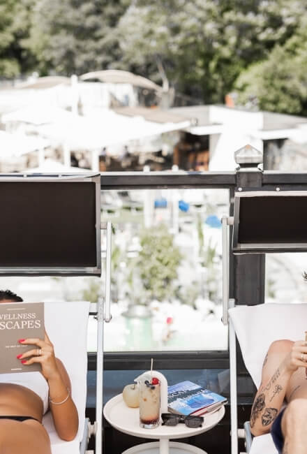 Persona su sdraio legge "WELLNESS ESCAPES" con drink e occhiali.