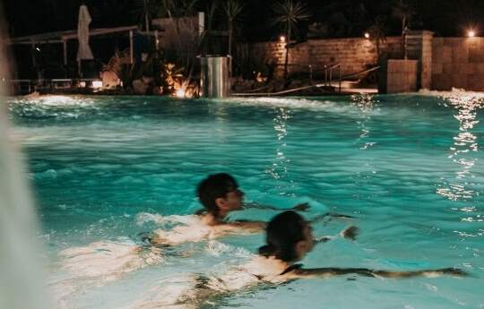 Due persone nuotano in una piscina illuminata di notte.