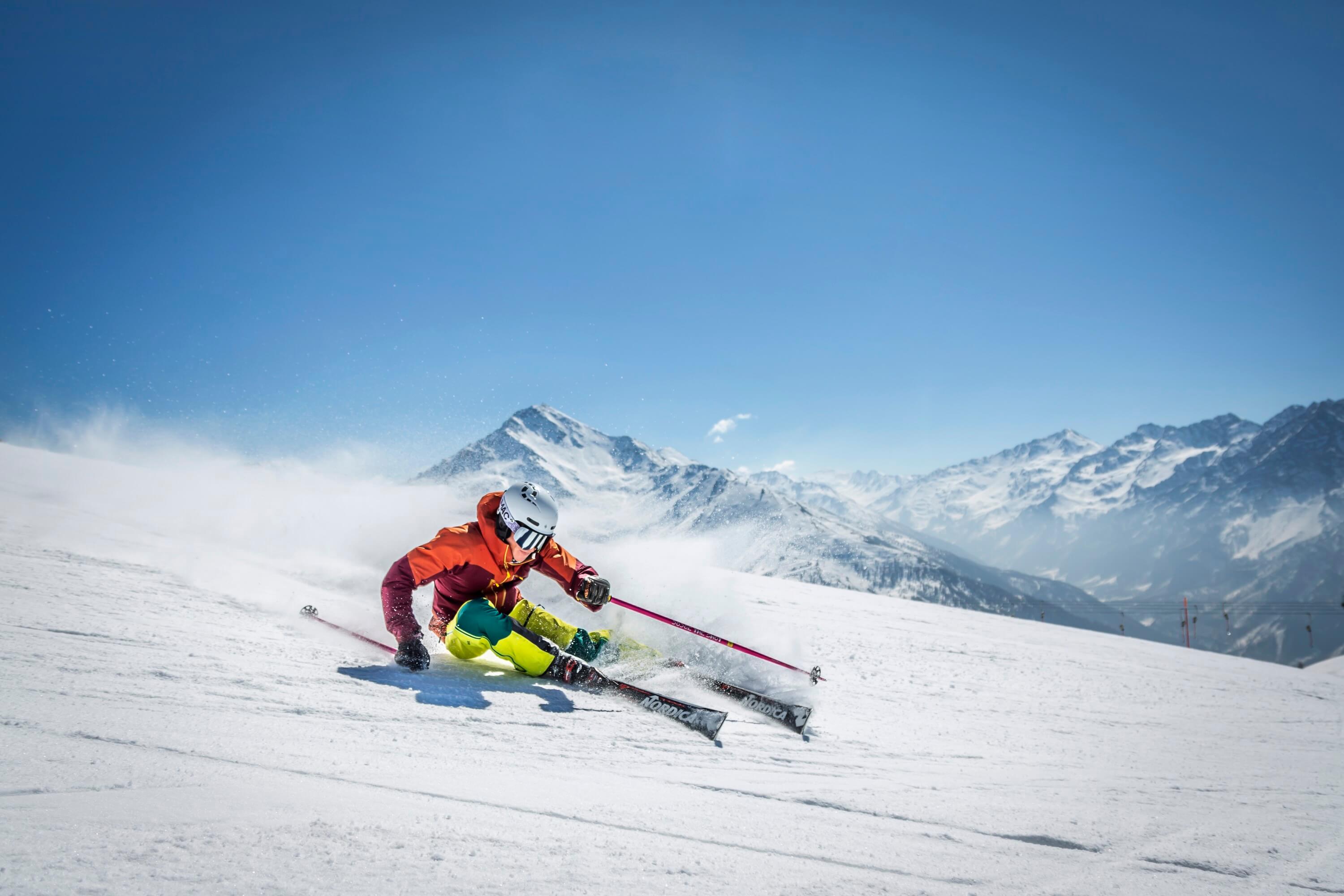 Skier in orange jacket and yellow pants carves down a snowy slope with mountains in the background under a blue sky.