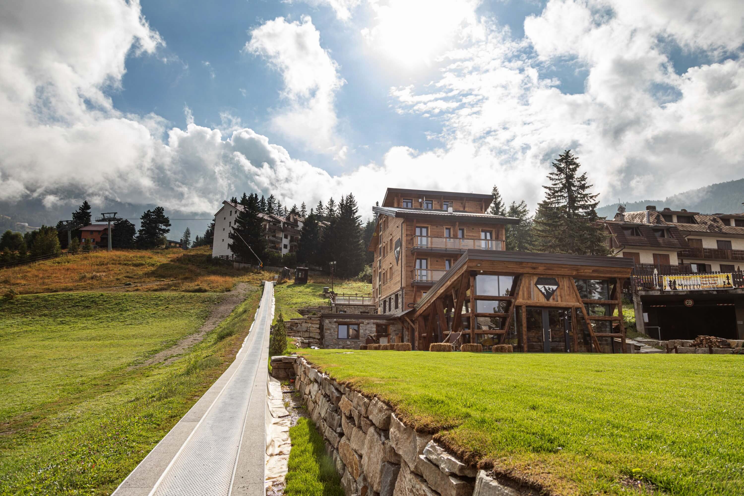 Serene mountainous landscape with a wooden building, lush greenery, and a bright blue sky with clouds.
