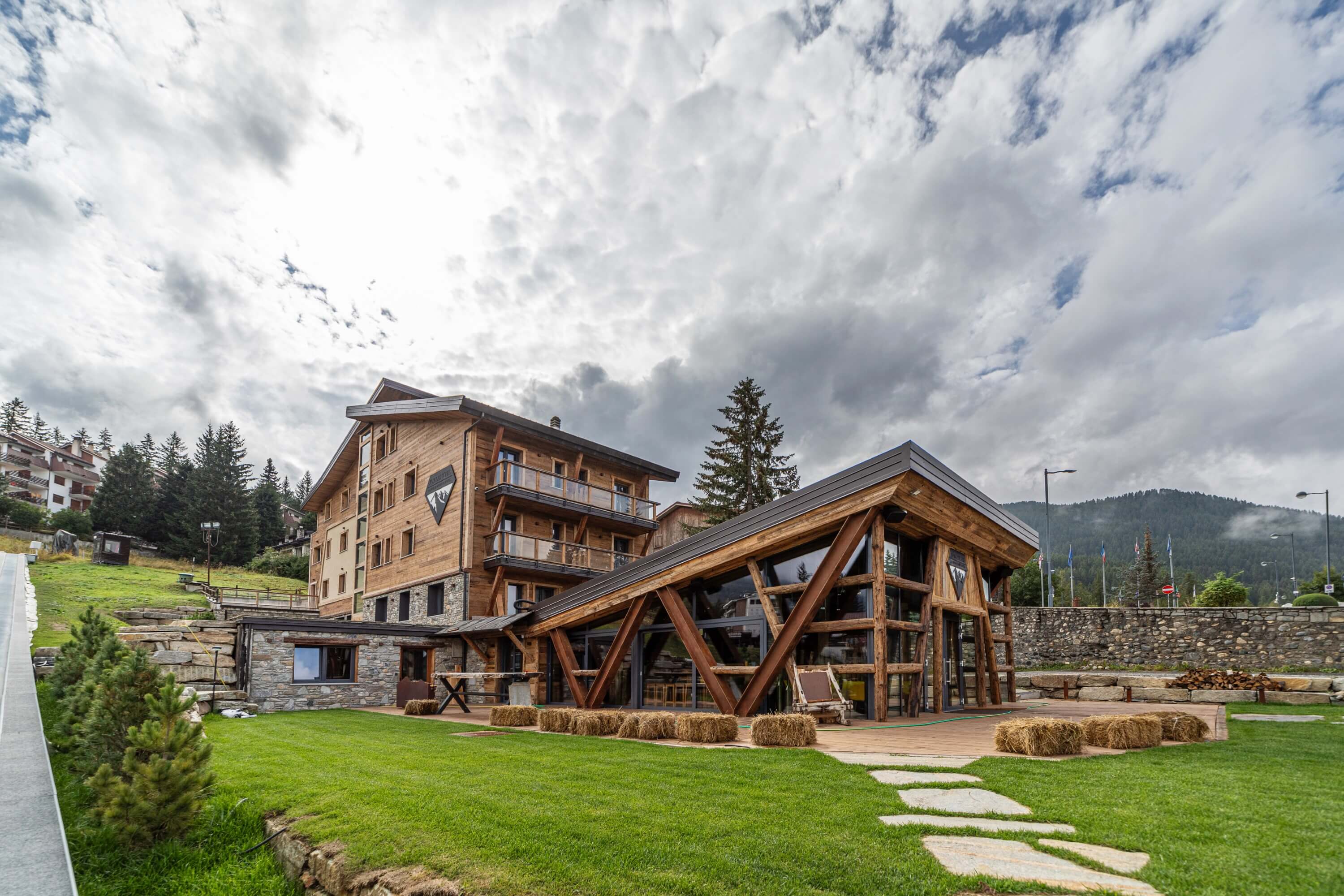 Multi-story wooden building with balconies, glass entrance, grassy area, trees, and mountains in a resort-like setting.