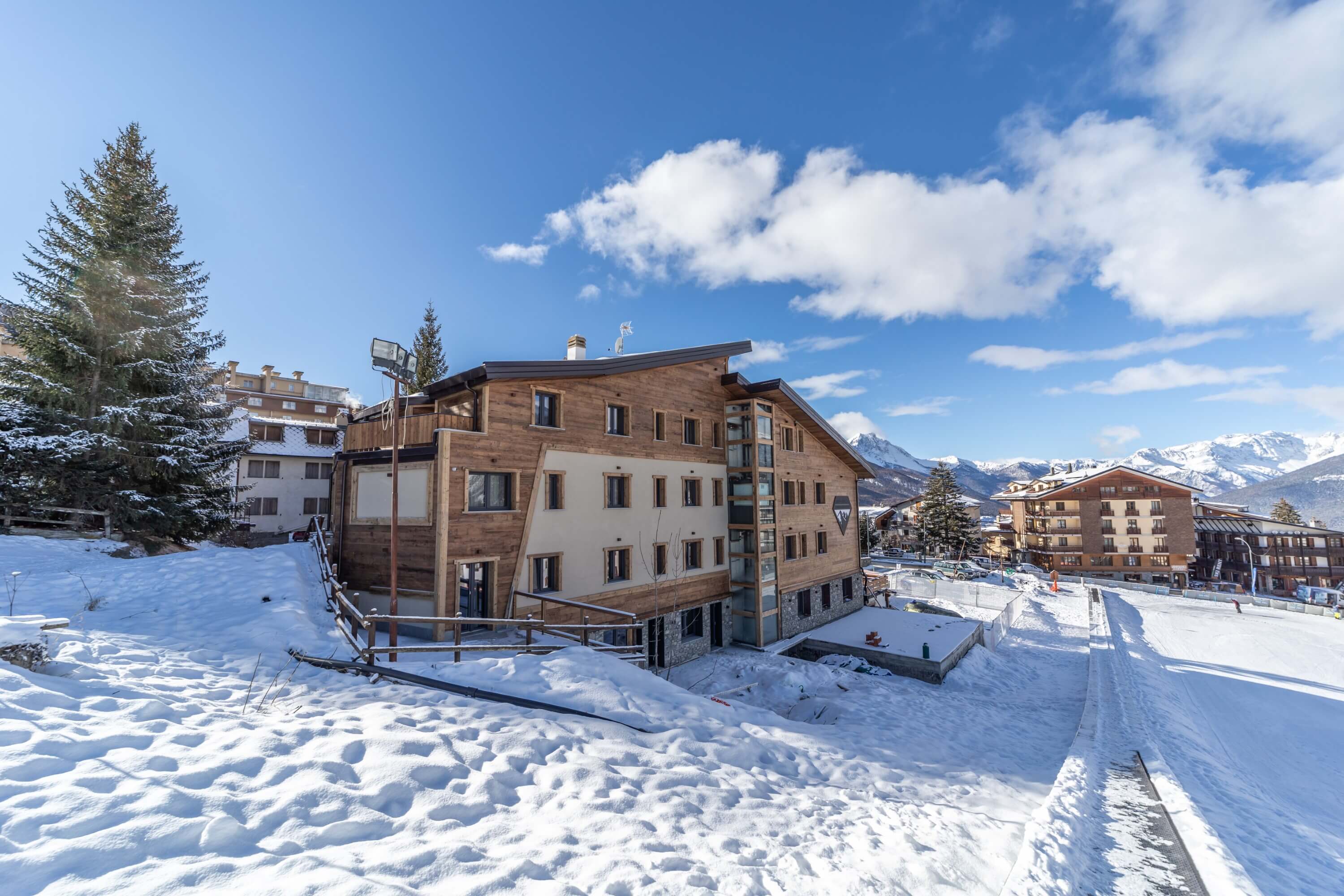 Serene winter scene featuring a unique building in snow, ski tracks, evergreen trees, and distant mountains under a blue sky.