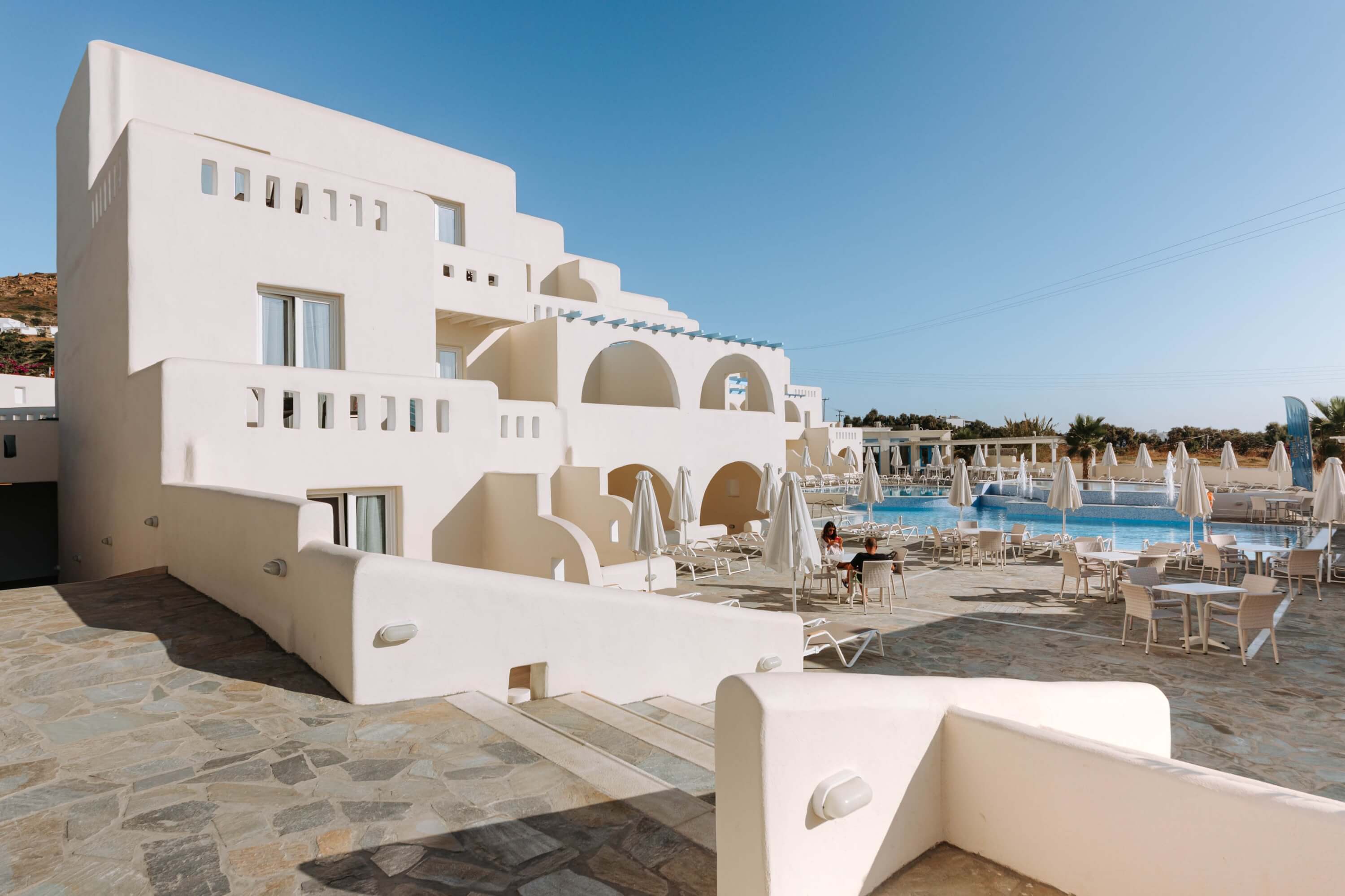 White multi-story building with a pool, patio, and two people seated at a table under umbrellas in a sunny resort setting.