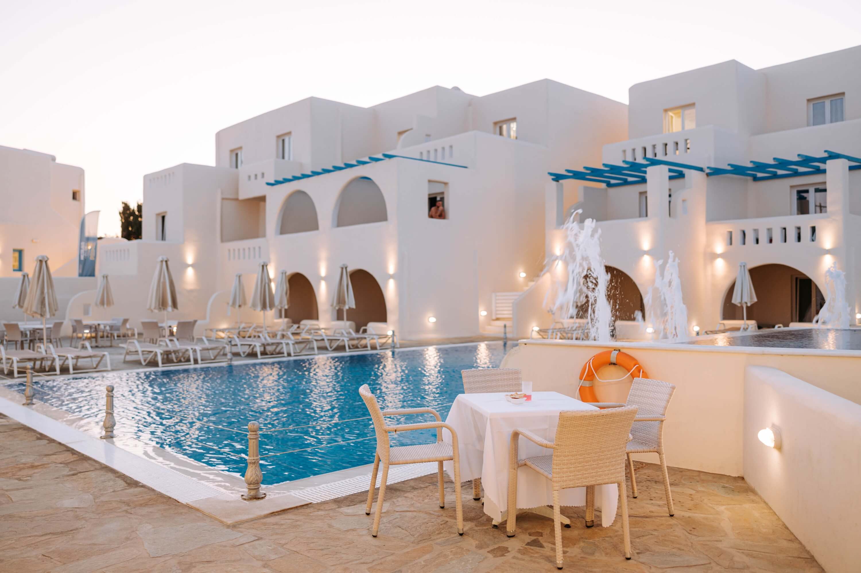 Serene hotel pool area with a large rectangular pool, circular pool, lounge chairs, and a table set for relaxation.