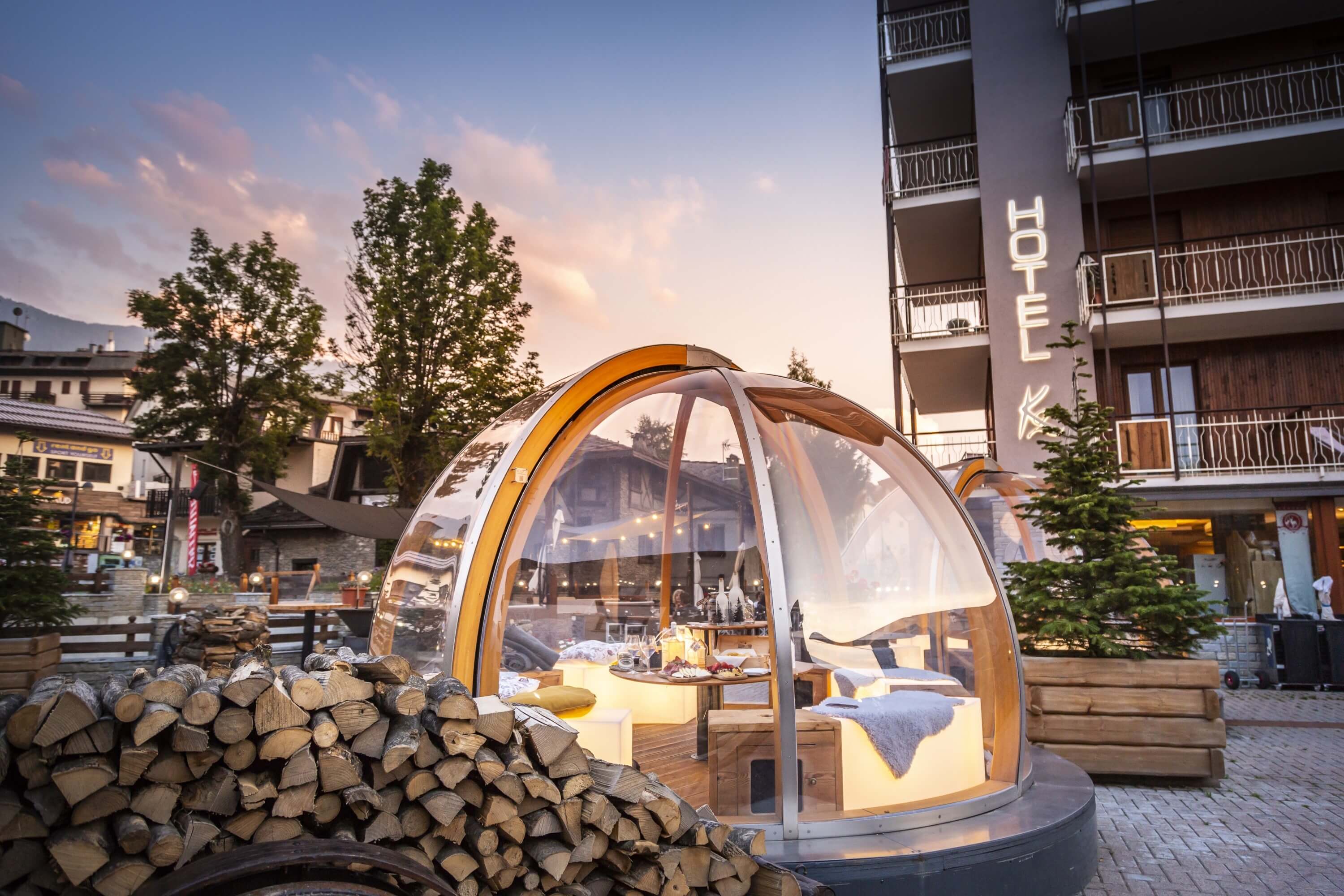 Hotel scene featuring a dome structure with a cozy interior, surrounded by trees and a tranquil sky at sunset.