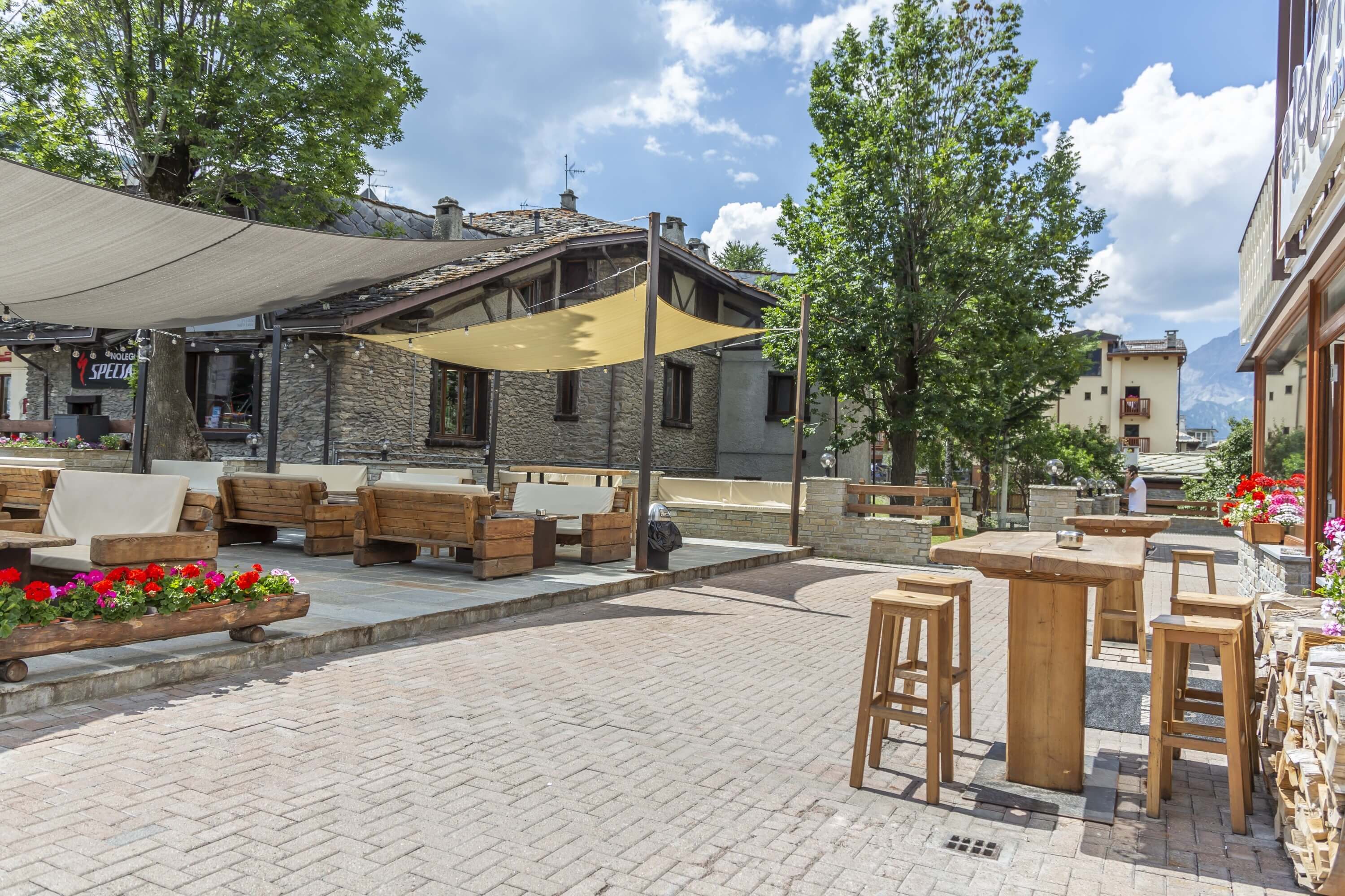 Outdoor seating area with wooden furniture, vibrant flowers, and a large tree, set against a picturesque town backdrop.