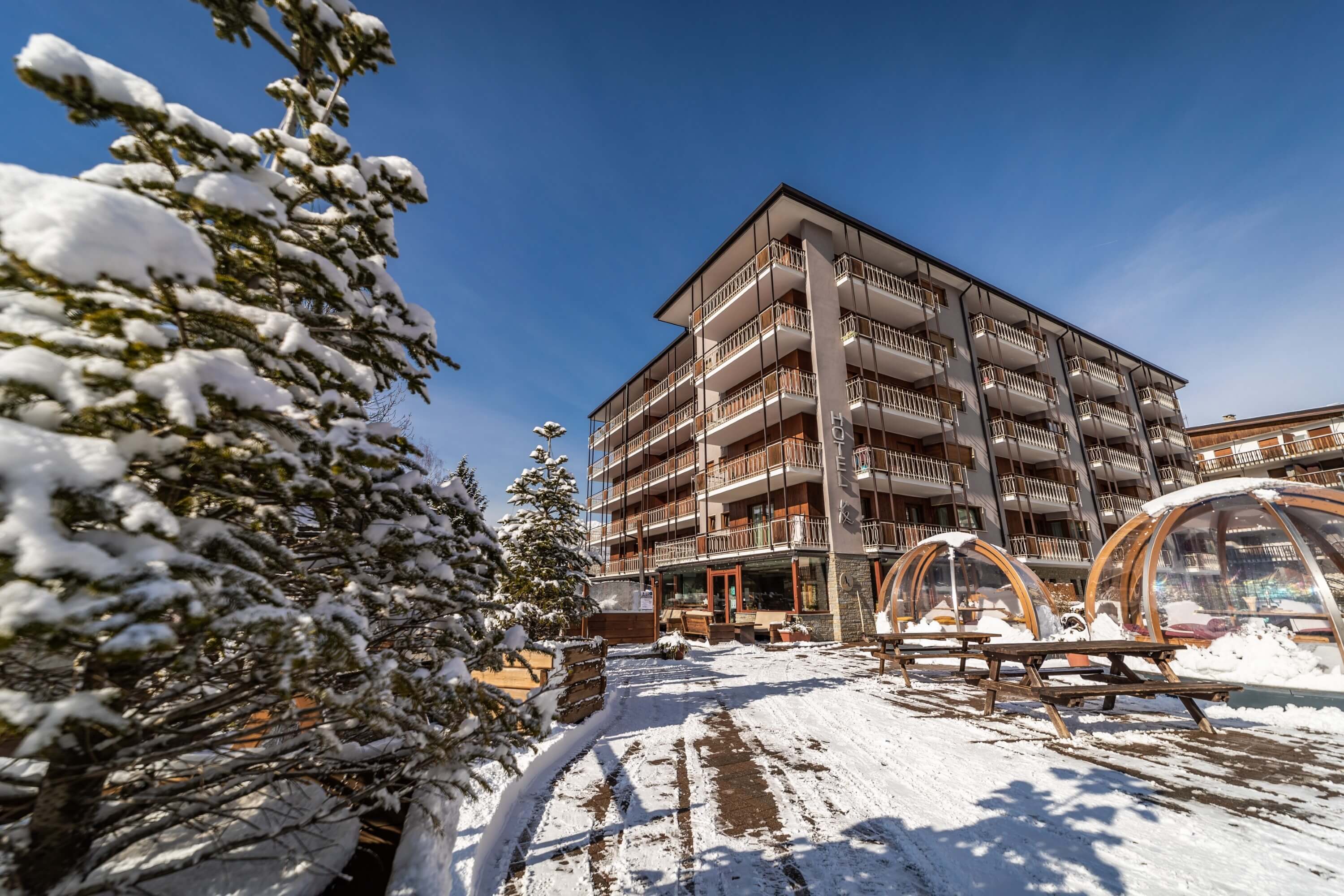 Multi-story hotel with balconies in a snowy landscape, featuring a sign "HOTEL AK" and transparent outdoor seating domes.