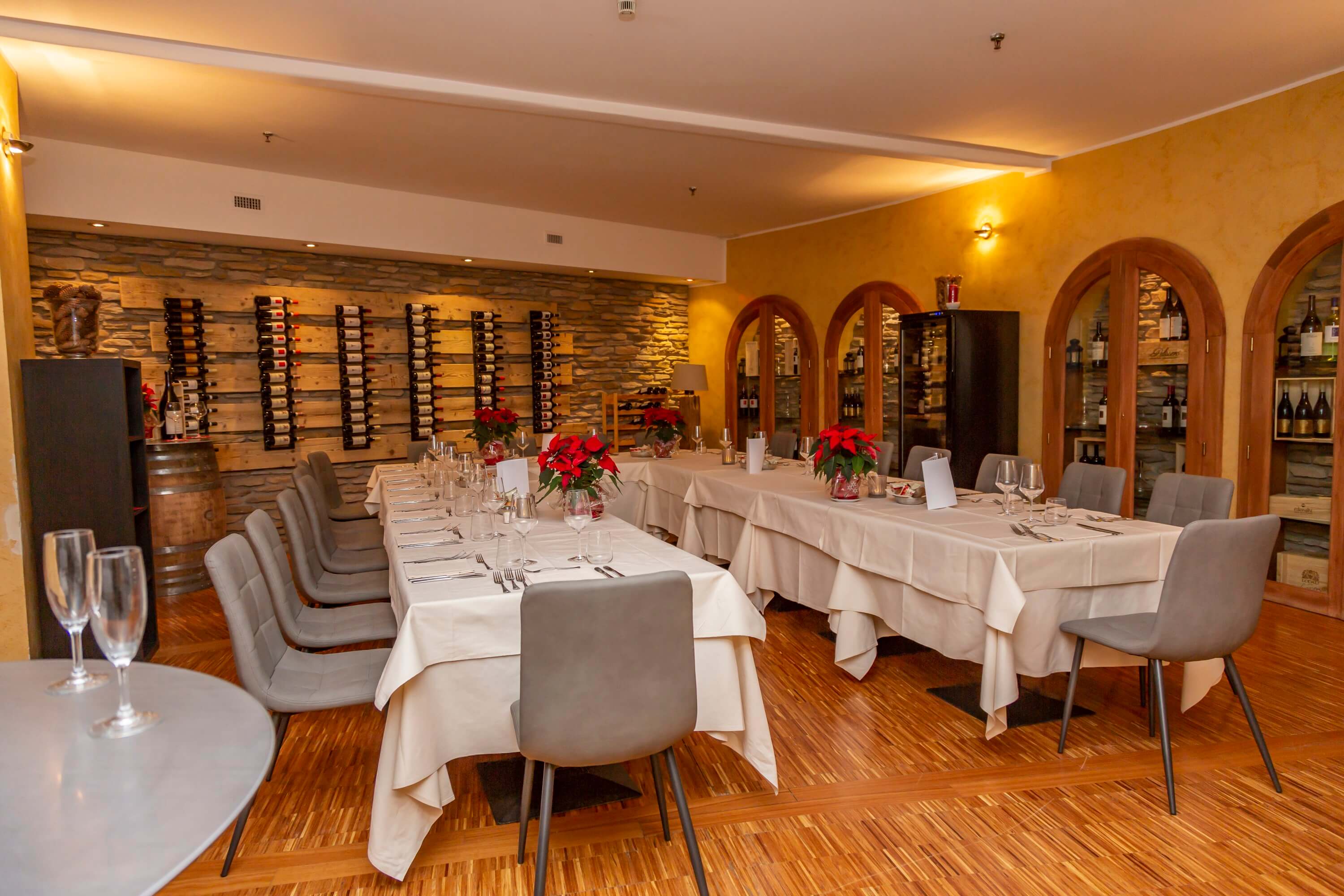 Cozy restaurant dining room with long tables set for dinner, red poinsettia centerpieces, and a wine wall in the background.