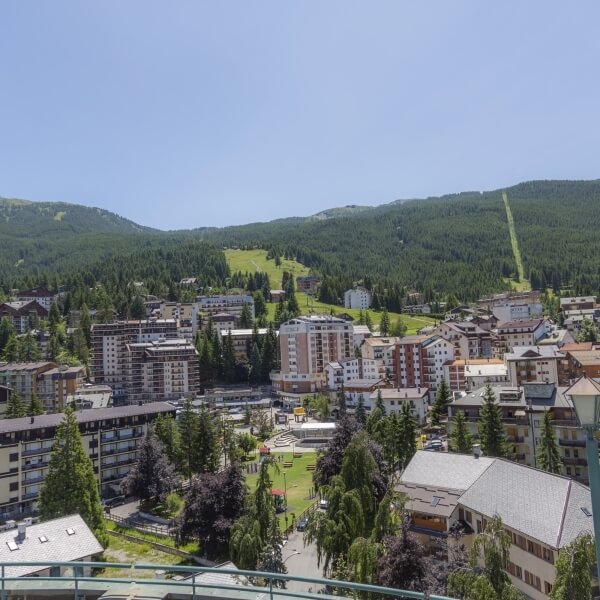 Vista panoramica di un piccolo paese montano con edifici bianchi, alberi, un'area verde centrale e montagne sullo sfondo.