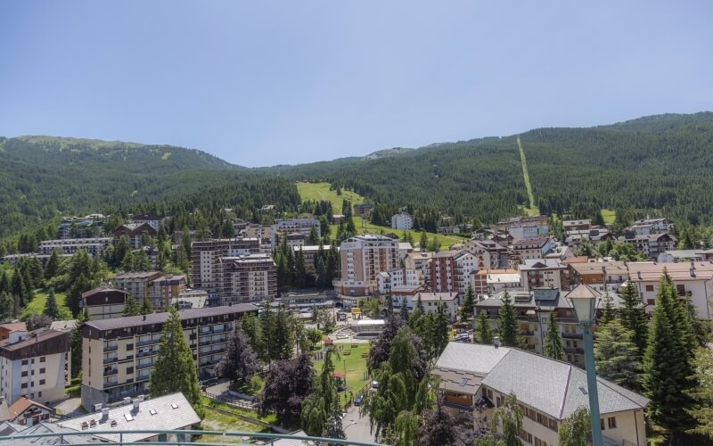 Molti edifici di stili architettonici misti circondati da alberi e vegetazione, con un cielo sereno e montagne sullo sfondo.