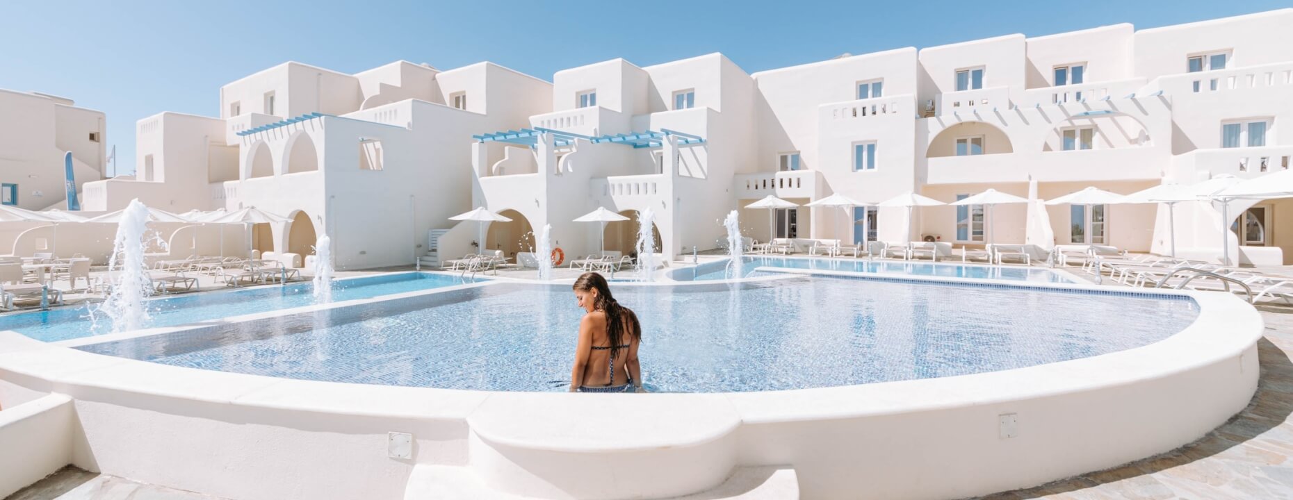 Una donna in bikini nero sta nella parte bassa di una piscina rettangolare circondata da edifici bianchi e lettini.