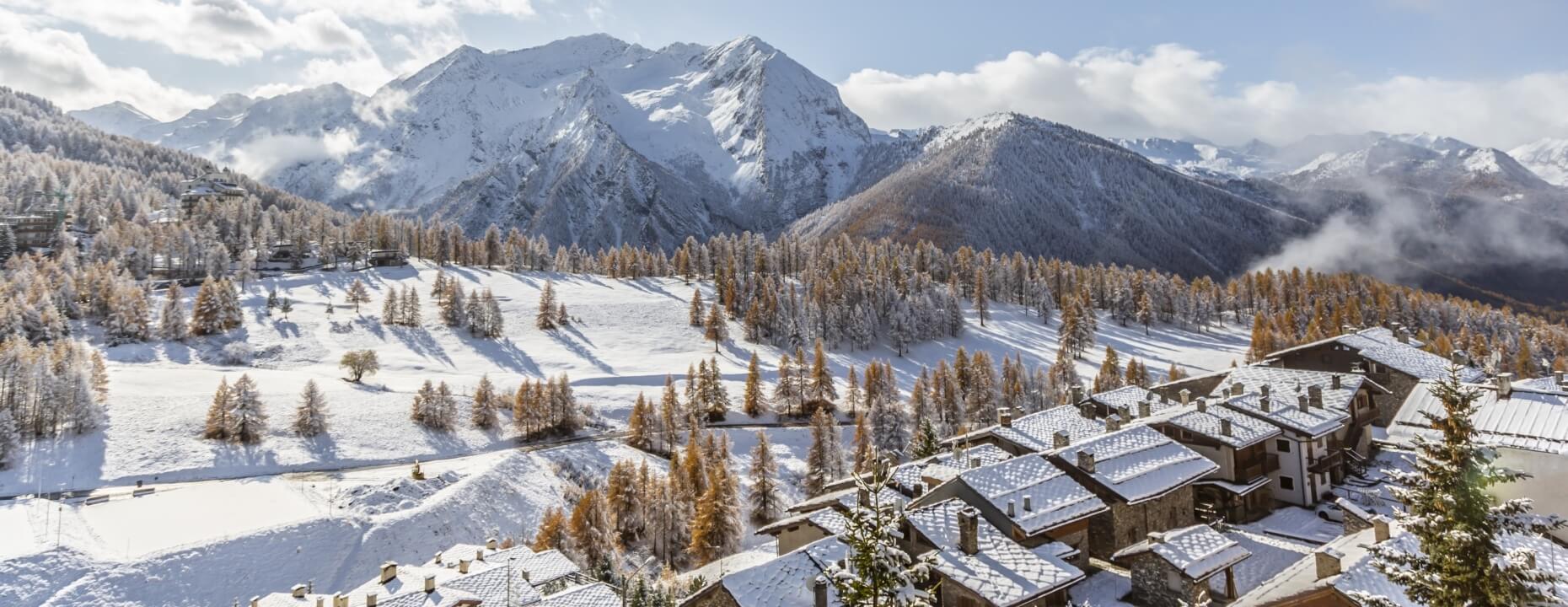 Serene winter landscape with snow-covered mountains, a village of snowy houses, and vibrant trees under a blue sky.