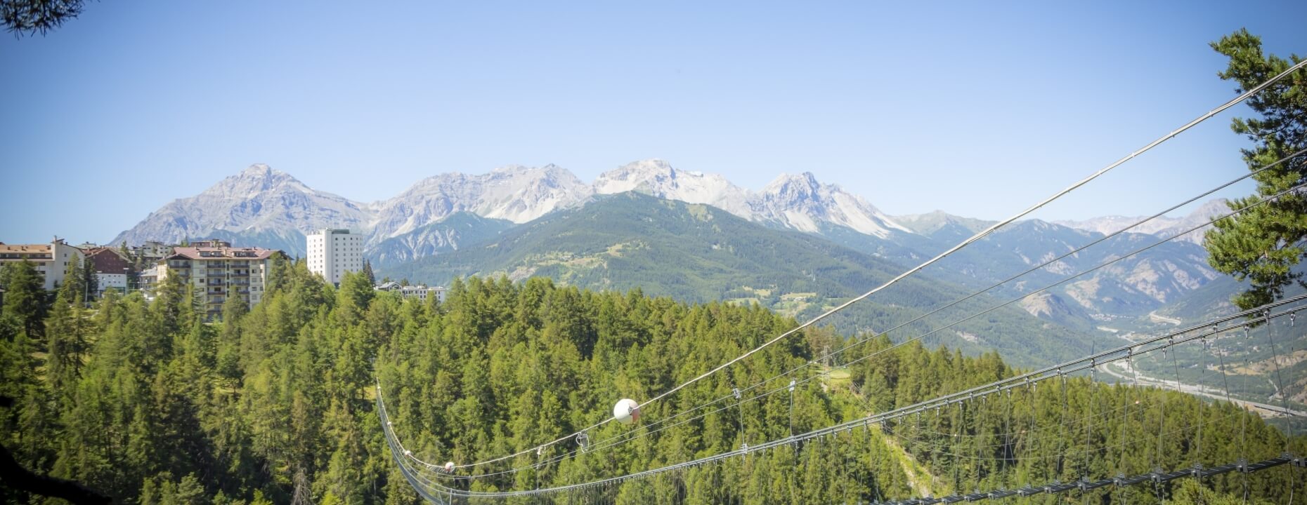 Scenic mountainous landscape with a suspension bridge, lush trees, buildings, and peaks under a clear blue sky.