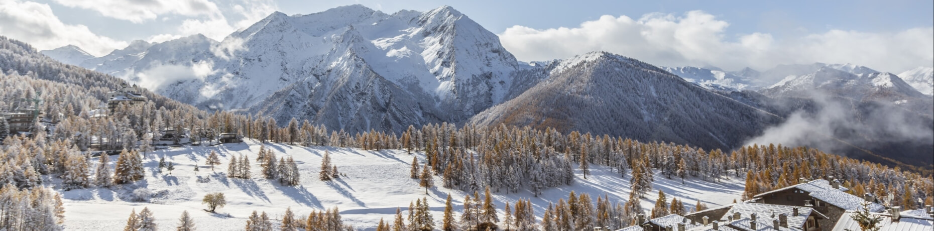 Panoramic view of snow-covered mountains, valleys, trees, and buildings under a bright, cloudy sky.