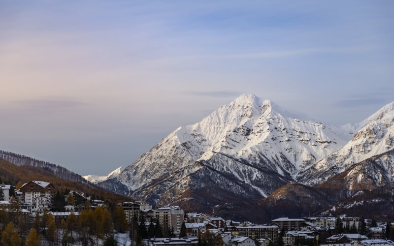 Un tranquillo paese di montagna ai piedi di una catena montuosa innevata, con edifici e alberi autunnali.