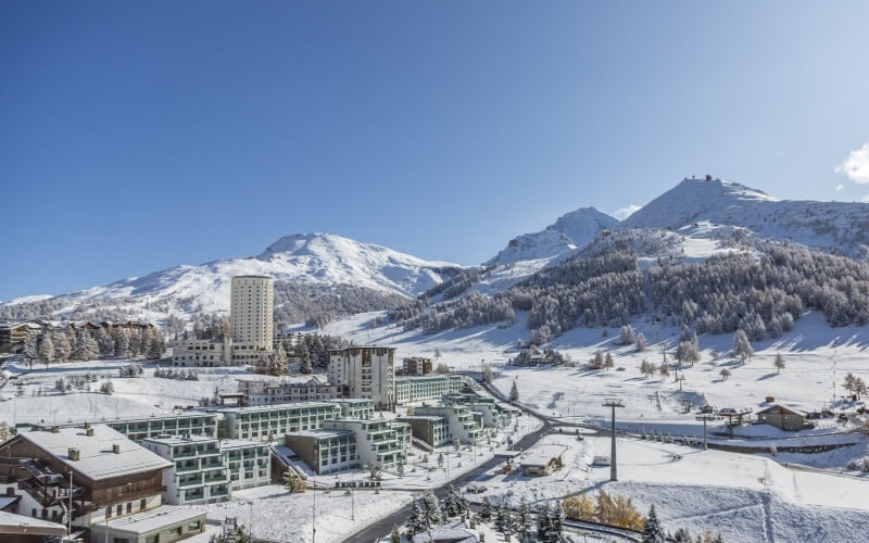 Winter scene of a mountainous town with snow-covered buildings, a winding road, and majestic mountains under a clear blue sky.