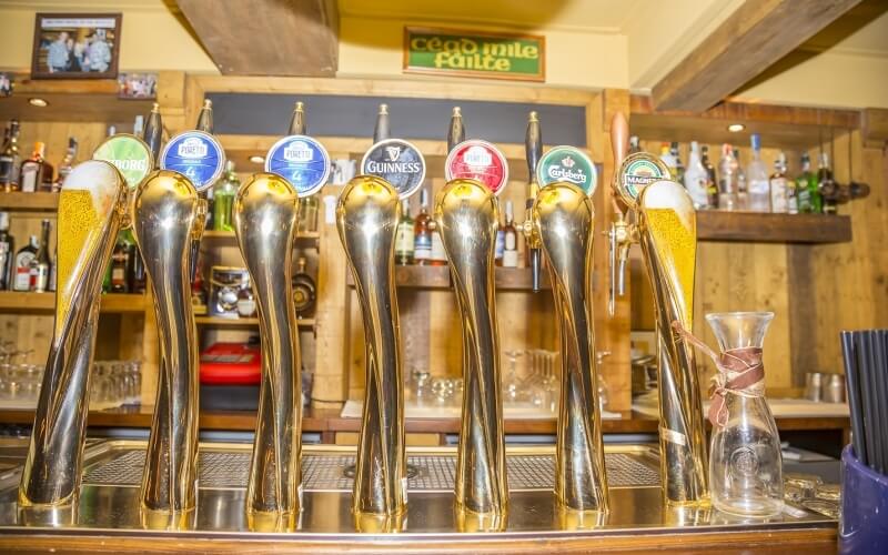 Stainless-steel bar top with nine beer taps, two filled glasses, and a carafe, set in a warm, inviting bar atmosphere.