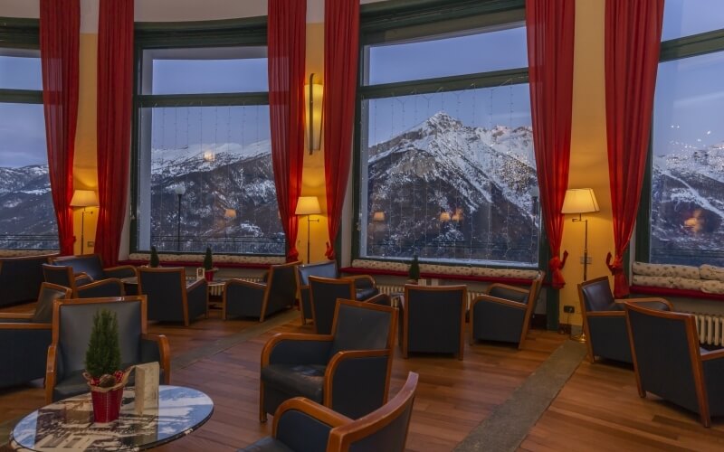 Lounge area with dark blue chairs, small tables, and mountain view, featuring warm lighting and yellow walls with red curtains.