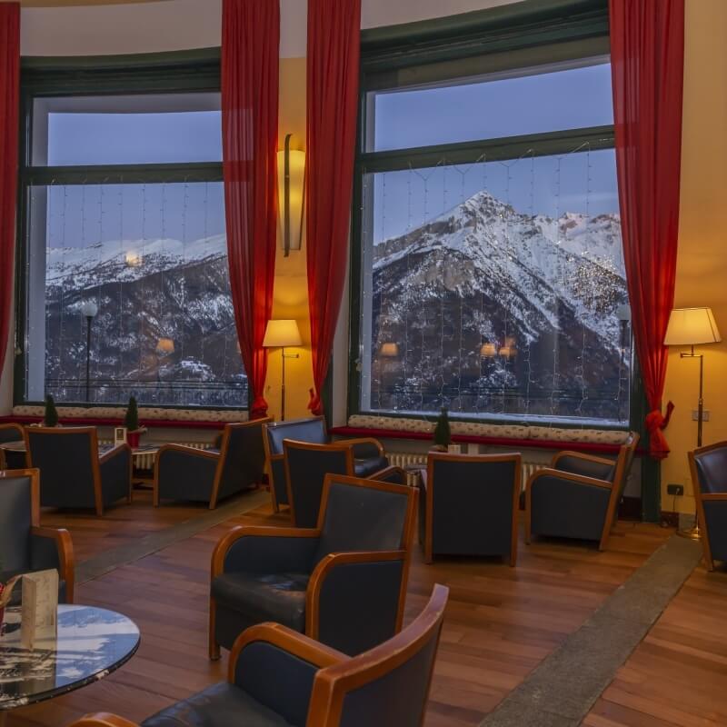 Cozy room with large windows, dark blue chairs, red curtains, and a view of snow-covered mountains under a blue sky.