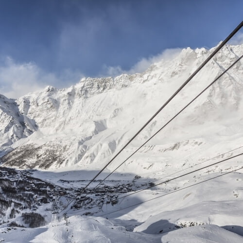 Snowy mountain range with ski lifts in the foreground under a blue sky with white clouds, creating depth and distance.