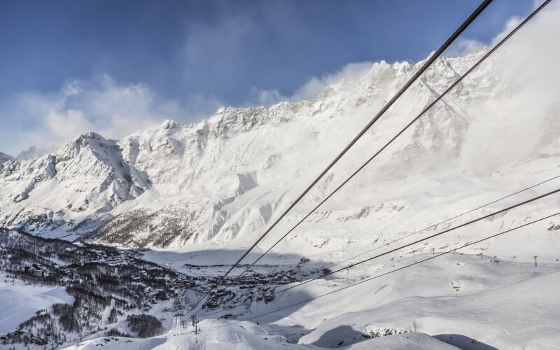 Winter scene featuring a ski lift, snow-covered slopes, and a misty mountain range under a blue sky with clouds.