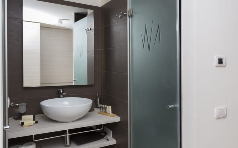 Bagno moderno con doccia walk-in, lavabo su piano grigio, grande specchio e piastrelle marroni scure. Atmosfera minimalista.